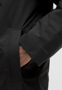 Hånd delvist inde i lommen på en sort Lululemon jakke, som viser ærmet med det diskrete præget brandlogo.