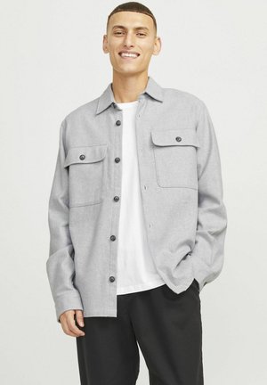Camisa gris de botón con tejido texturizado, que cuenta con dos bolsillos en el pecho y botones negros, llevada sobre una camiseta blanca. Corte relajado.