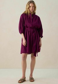 Robe courte violette avec un col haut, des manches bouffantes, une taille nouée et une silhouette fluide. Associée à des sandales à imprimé animal.
