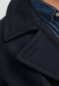 Collo di un cappotto di lana blu navy sovrapposto a quello di una camicia di denim con bottone visibile, concentrandosi sulla trama del tessuto e sul dettaglio dell'asola.