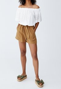Haut blanc à épaules dénudées avec détails en dentelle, short beige avec cordon de serrage, et sandales espadrilles vert olive avec accents de nœuds.