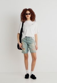 Νέα γυναίκα με σγουρά μαλλιά που φοράει μαύρα γυαλιά ηλίου, λευκό φαρδύ t-shirt, τζιν σορτς με σκισίματα, λευκές κάλτσες, μαύρα παπούτσια και μια μαύρη τσάντα ώμου.