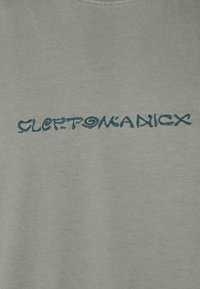 Light green cotton t-shirt featuring blue embroidered text "CLEPTOMANICX" in a stylized, swirly font across the chest area.