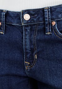 Jeans in denim blu scuro con cuciture gialle a contrasto, un bottone in rame e un'etichetta con il marchio in vita. Presenta passanti per cintura standard.