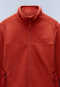 Giacca di pile rossa con colletto alto, zip completa e tessuto texture. Presenta tasche laterali e un logo a patch sul petto.