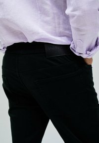 Jeans neri con una fascia elastica in vita, dotati di due tasche posteriori e una patch in cuoio con marchio in vita, abbinati a una camicia viola chiara.