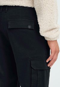 Schwarze Cargo-Hosen aus robustem Stoff, mit Seitentaschen und einem Knopfdetail, dazu ist ein weiches, cremefarbenes Fleece-Oberteil teilweise sichtbar.