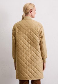 Femme blonde avec une queue de cheval portant un long manteau matelassé beige avec des poignets et un col côtelés, se tenant face à un mur blanc.