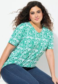 Zelené tričko s V-výstřihem a bílým botanickým potiskem, krátké rukávy, uvolněný střih, kombinováno s tmavě modrými džínami.