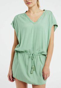 Robe courte vert menthe avec un décolleté en V, une taille à cordon de serrage et des manches courtes. Présente des accents en pompons sur le cordon pour un détail supplémentaire.
