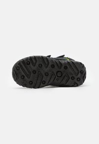 Semelle noire avec un motif de crampons multidirectionnels, présentant une surface texturée et des clous circulaires pour une adhérence accrue. Branding minimal visible.