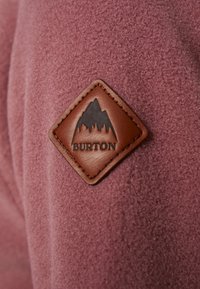 Écusson en cuir marron avec logo de montagne et texte "BURTON" sur tissu mauve texturé.