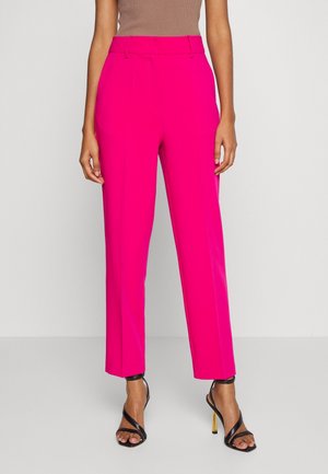 Vrouw draagt felroze op maat gemaakte broek en zwarte sandalen met hoge hakken en bandjes, staand tegen een effen achtergrond.