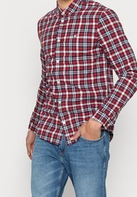 Röd och marinblå rutmönstrad långärmad skjorta med krage som knäpps ner och vita knappar, tillsammans med ljusblå jeans. Logotyp på bröstet.