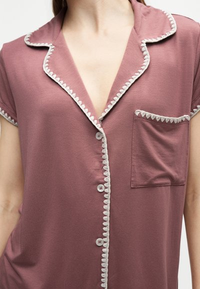 Chemise mauve à boutons avec bordure blanche en zigzag sur le col, le devant, la poche et les manches portée par une personne.