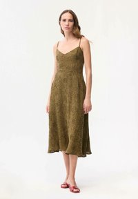 Woman wearing olive green floral midi dress with spaghetti straps and red open-toe flat sandals, standing against a plain light background.