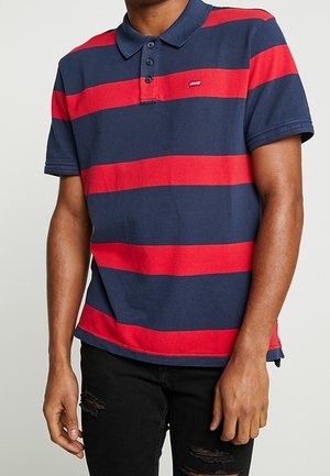 Mężczyzna ubrany w granatowo-czerwoną polo z poziomymi paskami i trzema guzikami oraz czarne, podarte jeansy.