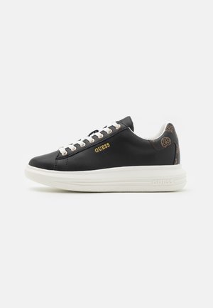 Zapatillas de cuero negro con suela de goma blanca, logotipo dorado y cordones blancos. Acento de tela detallado en el talón con un patrón marrón.
