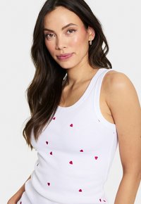 Camiseta sin mangas blanca de canalé con pequeños patrones de corazones rojos dispersos, con un escote redondo y tirantes anchos.