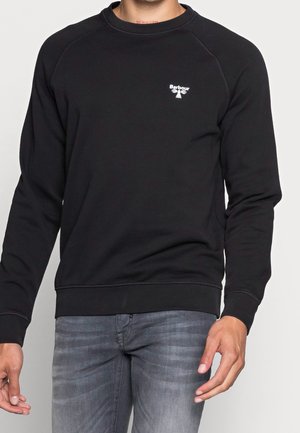 Mężczyzna w czarnej, długorękawowej bluzie Barbour i wyblakłych szarych jeansach, stojący z rękami swobodnie zwisającymi po bokach.