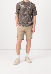 T-shirt camouflage dans des tons terreux, associé à un short cargo beige avec des poches latérales, et des chaussettes blanches avec des baskets beiges.