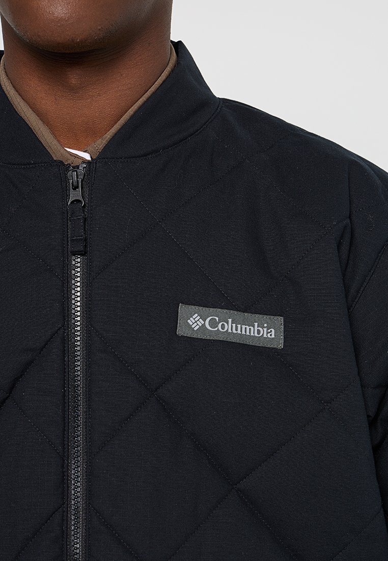 Czarna pikowana kurtka z wysokim kołnierzem i zapięciem na zamek. Posiada szary naszywkę logo Columbia na lewej piersi. Teksturowany materiał, minimalistyczny design.