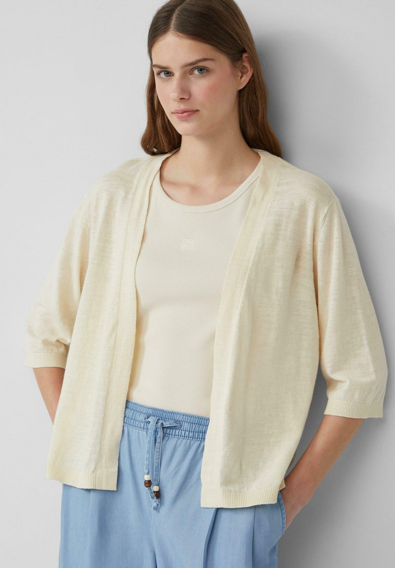 Cardigan leggero e cremoso beige con maniche a trequarti, apertura frontale e dettagli a coste, indossato sopra una canotta beige e pantaloni blu.