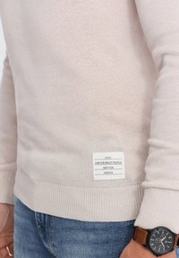 Pull en maille beige clair avec un tissu texturé, ourlet côtelé et une étiquette affichant 'Votez pour les bonnes personnes, pas pour XXXXXX.'