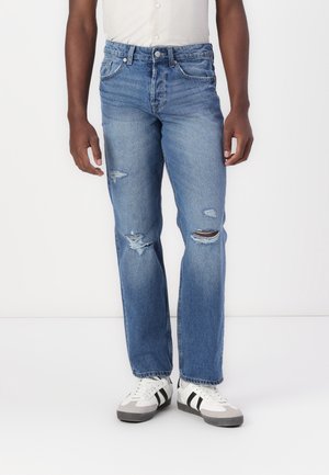 Man wearing blue ripped jeans and white sneakers with black stripes, standing against a plain light background.