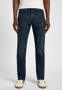 Jeans in denim scuro con taglio dritto, dotati di cinque tasche, cuciture sottili e accessori in metallo, abbinati a sneakers bianche.