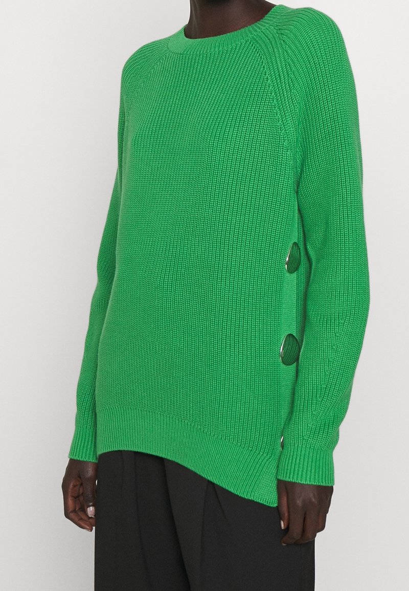Grüner Strickpullover mit Rundhalsausschnitt, der über eine gerippte Textur, seitliche Knopfdetails und einen leicht verlängerten Saum verfügt.