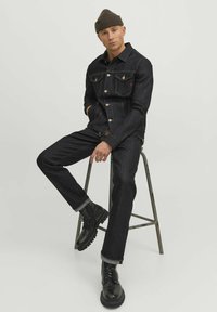 Veste en denim foncé avec des boutons en ton cuivré et deux poches poitrine, associée à un jean foncé assorti. Bottes à lacets noires et bonnet marron.