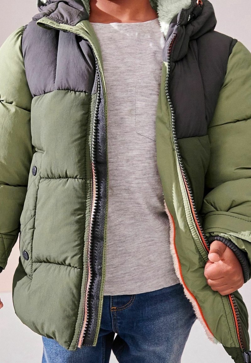 Chaqueta acolchada con capucha, que presenta una combinación de colores en verde oscuro y gris, cierre de cremallera y detalles de botones. Lleva puesta sobre una camiseta gris.