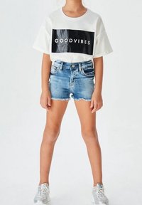 T-shirt bianca in cotone con la scritta “GOODVIBES” in nero, abbinata a pantaloni shorts in denim strappati e sneakers argentate, in piedi contro uno sfondo neutro.
