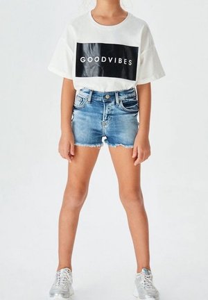 Vit bomull t-shirt med "GOODVIBES" i svart text, i kombination med slitna denimshorts och silverfärgade sneakers, stående mot en neutral bakgrund.