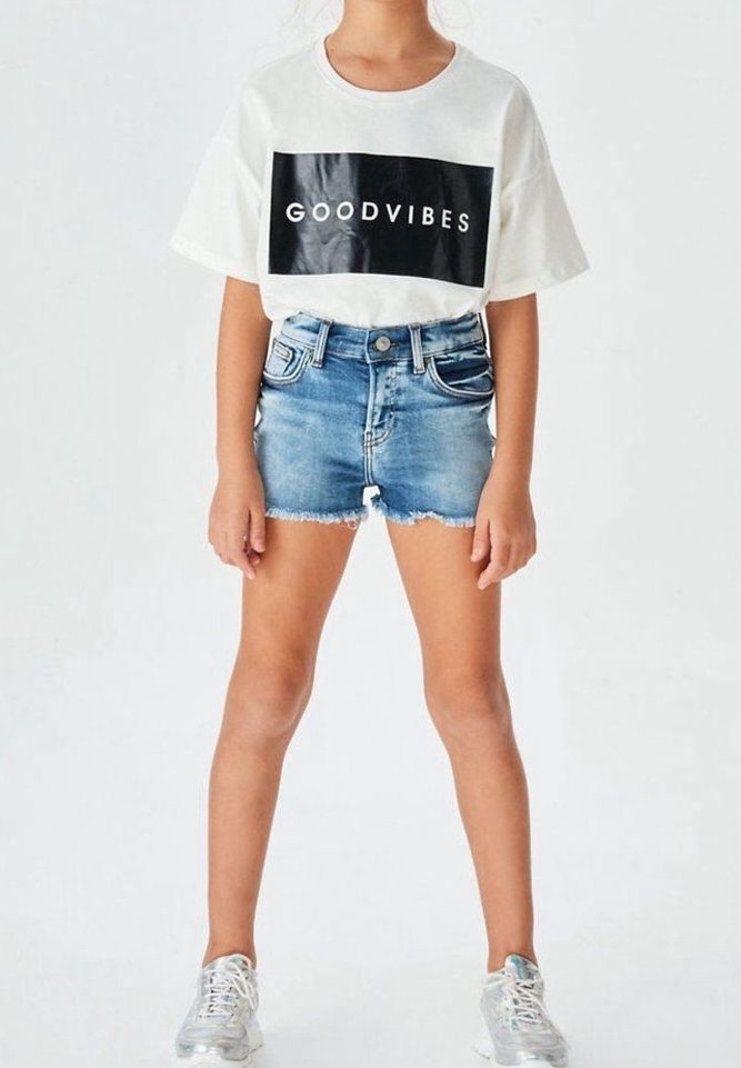 T-shirt bianca in cotone con la scritta “GOODVIBES” in nero, abbinata a pantaloni shorts in denim strappati e sneakers argentate, in piedi contro uno sfondo neutro.