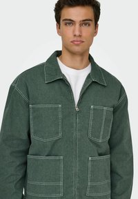 Chaqueta de mezclilla verde con cierre frontal, dos bolsillos en el pecho y dos bolsillos inferiores, con costuras blancas en contraste y cuello.