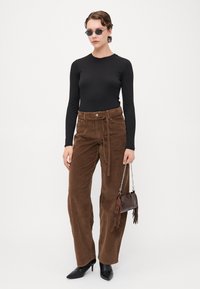 Pantaloni largi din corduroy maro, cu o talie legată, purtați cu un top negru cu mânecă lungă și tocuri negre ascuțite. Accesoriile includ o mică geantă maro.