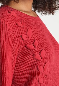 Punainen neulottu villapaita, jossa on resorikuvio ja lehtimuotoiset applikaatiot olkapään kohdalla. Pyöreä pääntie, pitkät hihat ja rento istuvuus.