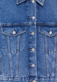 Close-up van de voorkant van een blauwe spijkerjack met metalen drukknoopjes en twee borstzakken met kleppen en knoopsluitingen.