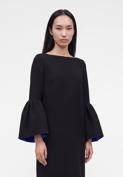 Γυναίκα με μακριά μαύρα μαλλιά που φοράει μαύρο φόρεμα με φαρδιά μανίκια καμπάνα και μπλε εσωτερική επένδυση, στέκεται μπροστά σε απλό φωτεινό φόντο.