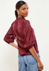 Femme aux cheveux bruns attachés, portant des boucles d'oreilles en or, un blouse bourgogne à texture avec des manches bouffantes, et un jean bleu clair, se retournant par-dessus son épaule.