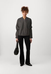 Casaco bomber cinza com fecho à frente, colarinho canelado e mangas balão, combinado com calças pretas estilo flare e uma mala preta.