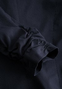 Granatowy materiał w drobne białe kropki, z marszczonym mankietem. Gładka tekstura z miękkim układem i subtelnym połyskiem.