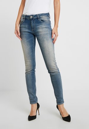 Jeans ajustados de mezclilla azul deslavada con una cintura media, diseño de cinco bolsillos y detalles en metal plateado, combinados con tacones altos negros.