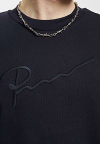 Svart hettegenser med en teksturert, tonalt "RM"-logo og et glattet, kontrastfarget linjemønster. Akkompagnert av et sølvfarget kjede.