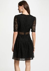 Abito in pizzo nero con maniche corte a ondine, corpino aderente e gonna svasata. Presenta un motivo testurizzato e un dettaglio sul retro aperto.