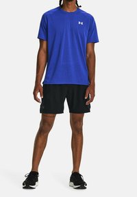 Modré sportovní tričko s krátkými rukávy, kulatým výstřihem a logem na hrudi, v kombinaci s černými šortkami a černými běžeckými botami.