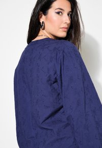 Top blu navy a maniche lunghe con ricami floreali a rilievo, scollatura tonda e vestibilità morbida, caratterizzato da una vista posteriore e maniche leggermente puff.