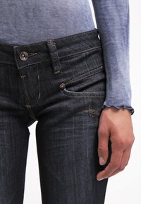 Jeans de mezclilla azul oscuro con una textura suave, cierre de botón de cobre, cinco bolsillos y detalles de costura visibles. Acompañados de una blusa azul clara.